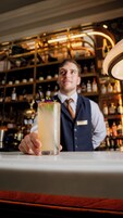 Un barman tient un cocktail décoré d’une garniture, dans un bar où l’on aperçoit, à l’arrière-plan, des étagères remplies de bouteilles.