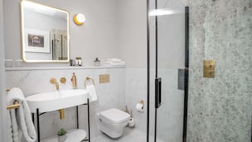 Un baño moderno con lavamanos blanco, detalles dorados, cabina de ducha con mampara de vidrio y paredes de mármol.