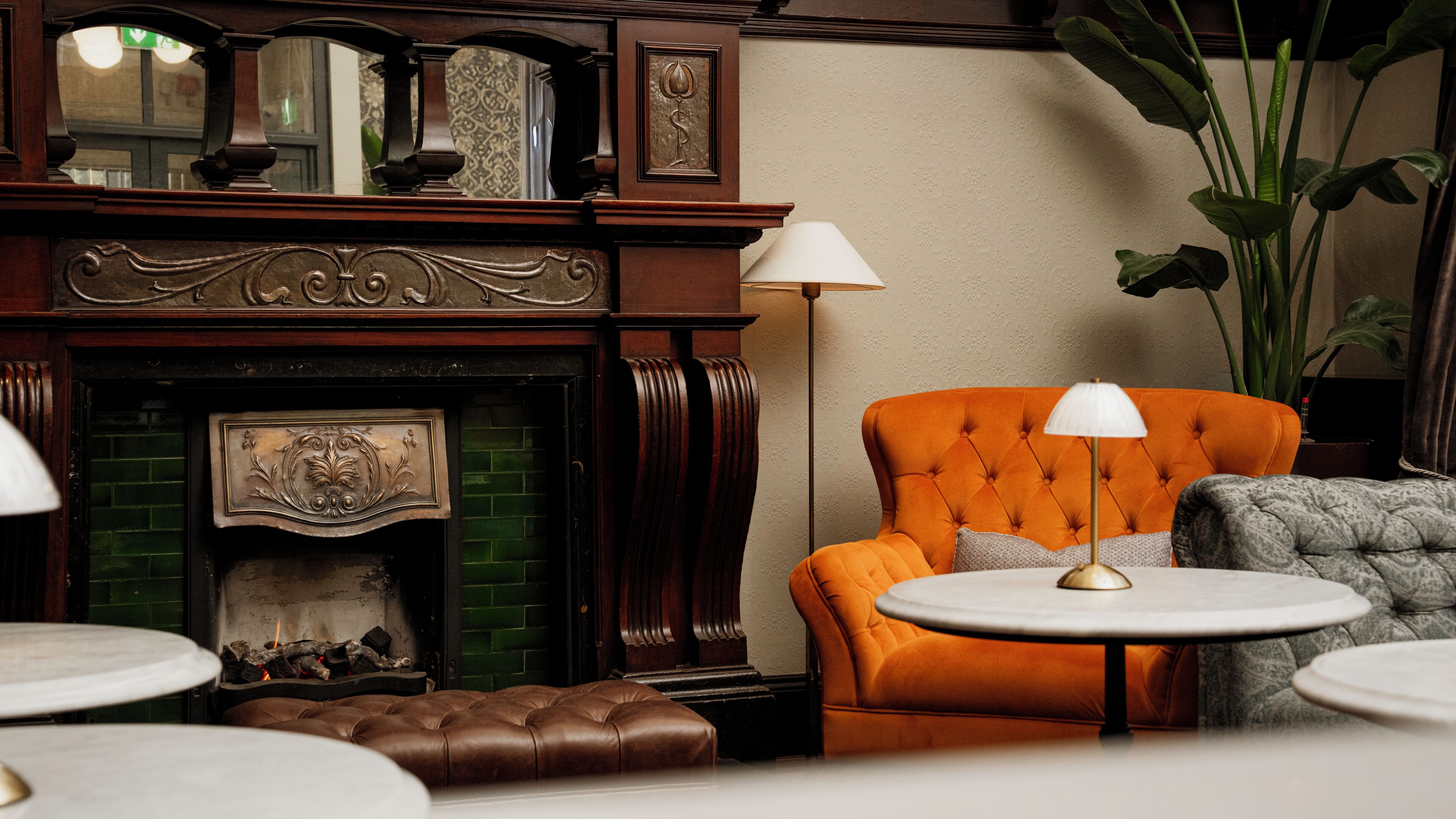 Una habitación con chimenea, un sillón naranja con capitoné, un sillón gris y una mesa redonda con lámpara.
