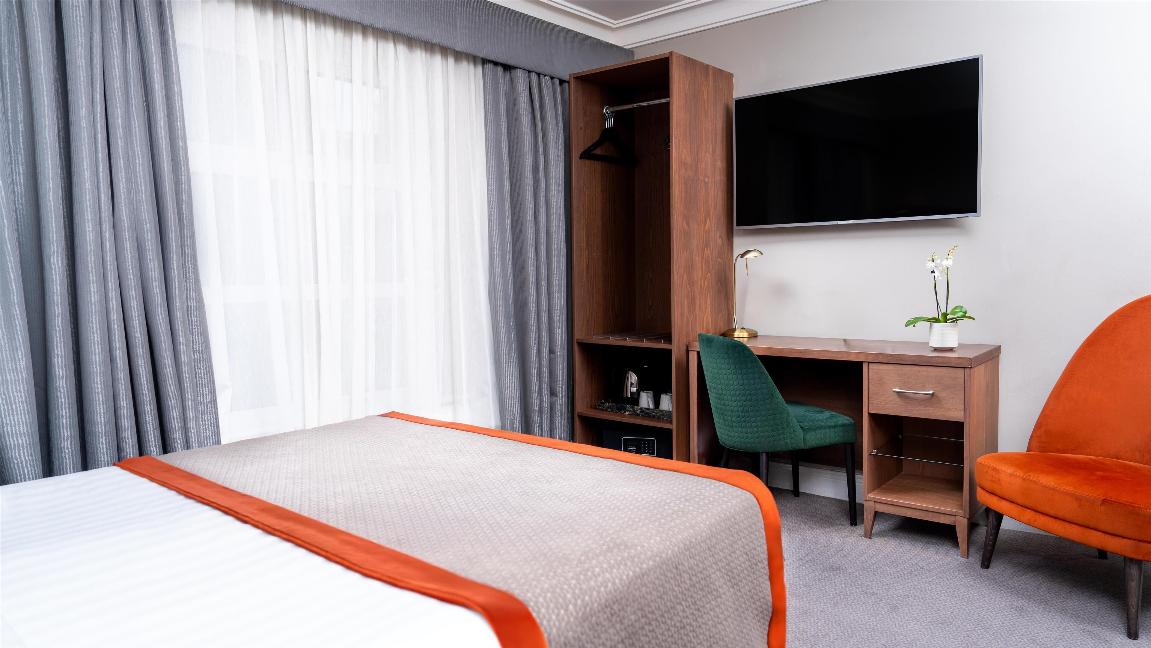 Una habitación de hotel con cama, escritorio con silla, televisor y una planta.