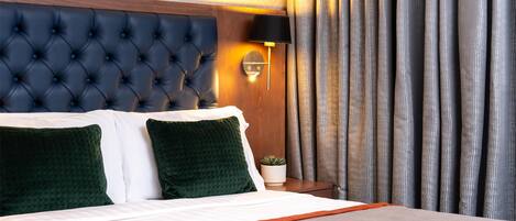 A hotel room with a tufted headboard, a bedside lamp, a potted plant, and a view of the outside through curtains.