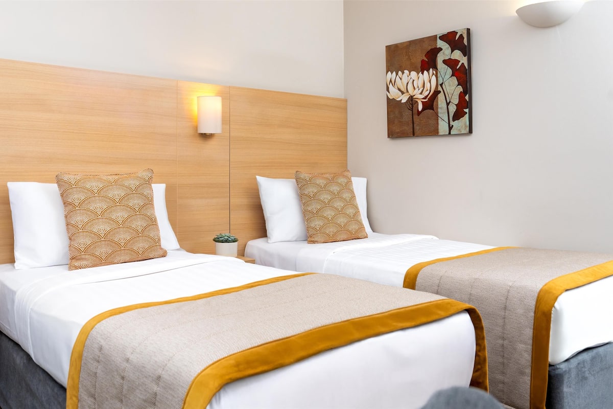 Habitación de hotel con dos camas, cada una con cabecera, ropa de cama blanca y un detalle amarillo. Hay lámparas de pared y una pequeña planta en maceta sobre la mesita de noche.