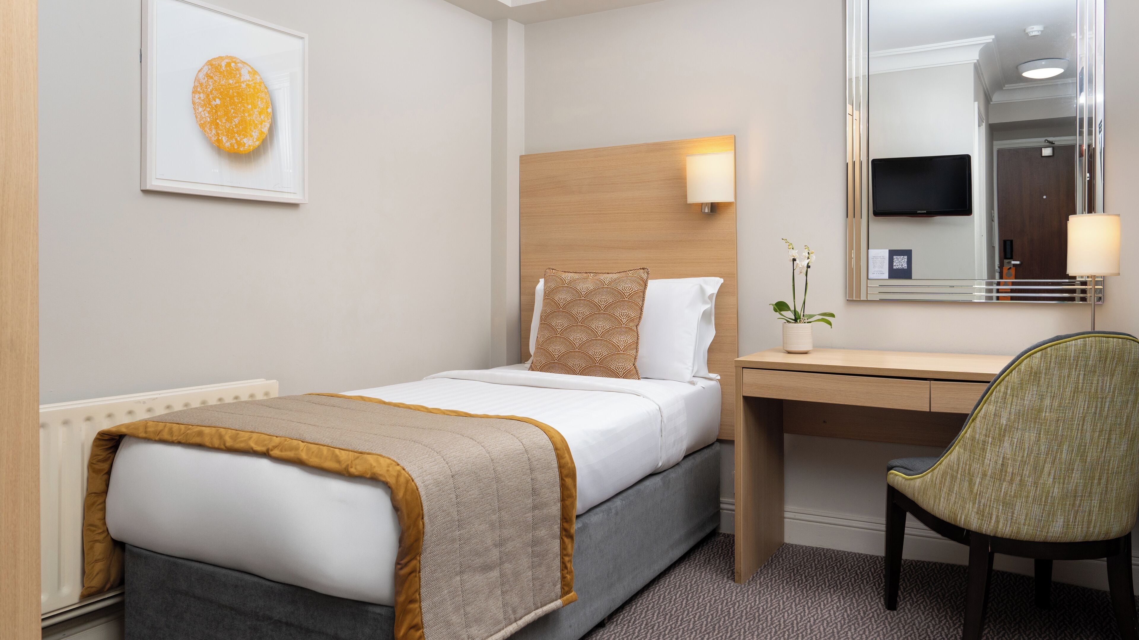 A hotel room with a bed, a desk, a chair, a bedside table, a lamp, a mirror, and a framed picture on the wall.