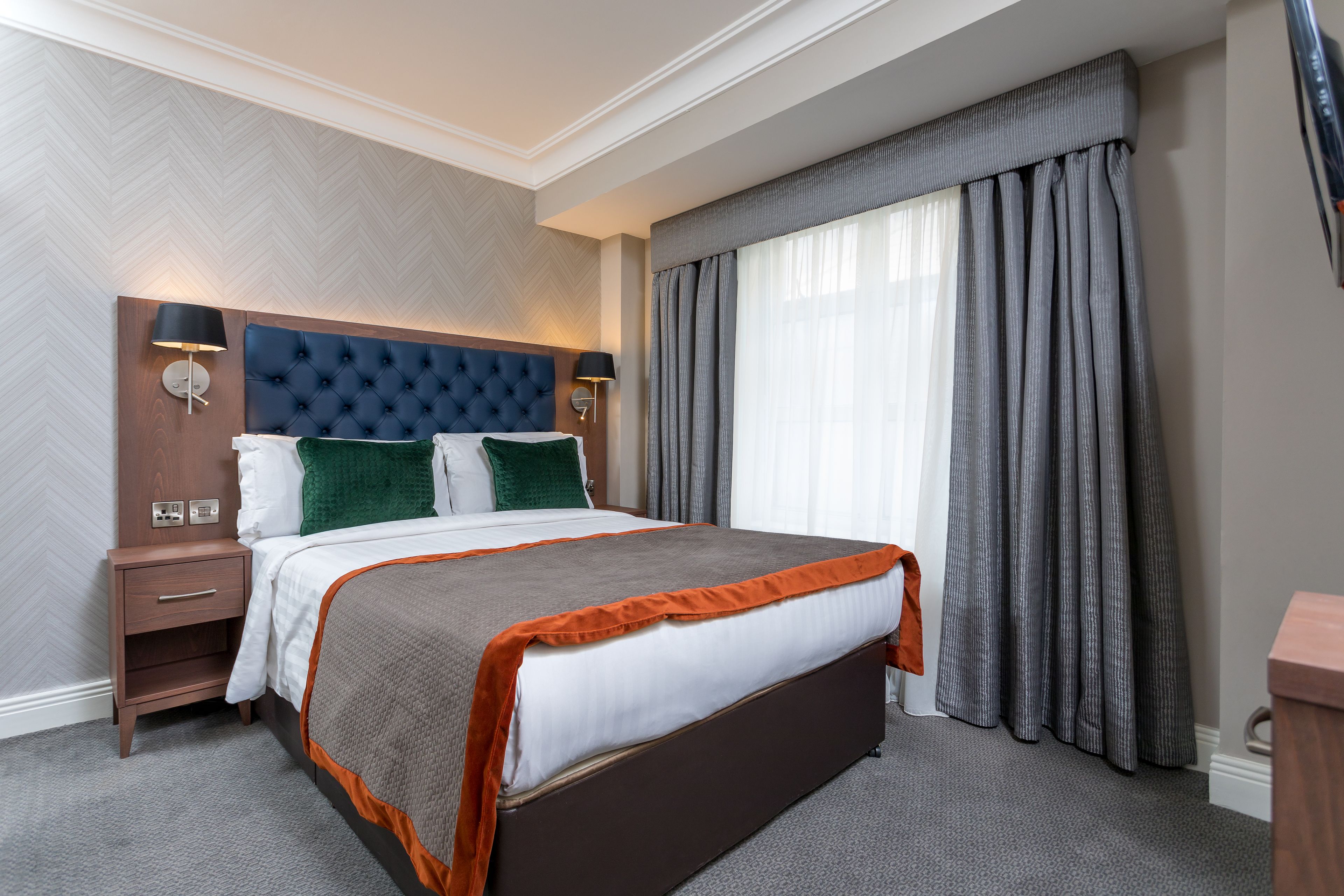 Una habitación de hotel con una cama grande, una mesita de noche, una lámpara de pared y un ventanal con cortinas.
