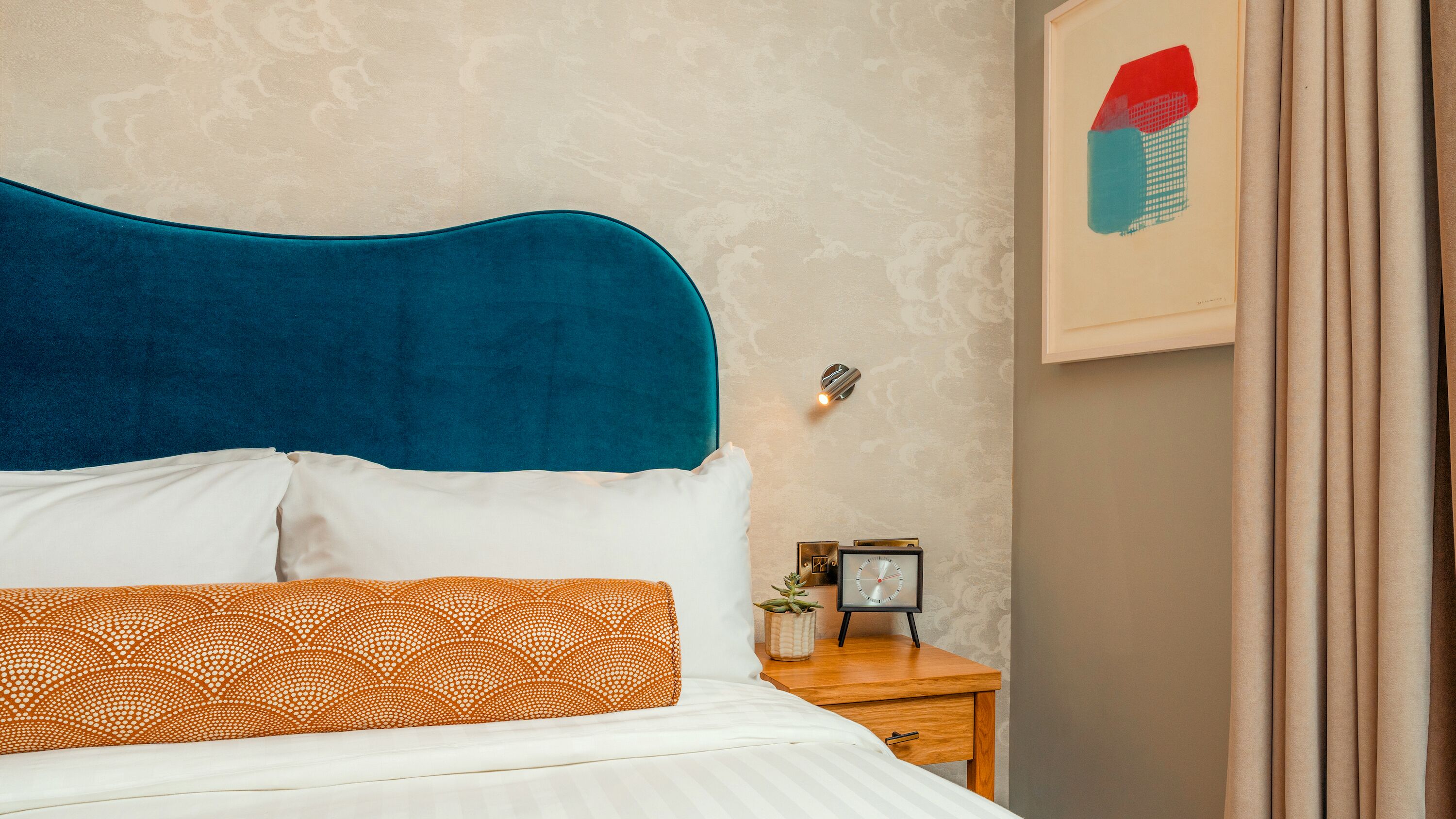A neatly made bed with a blue headboard, white and orange patterned pillows, and a small bedside table with a clock and a potted plant.