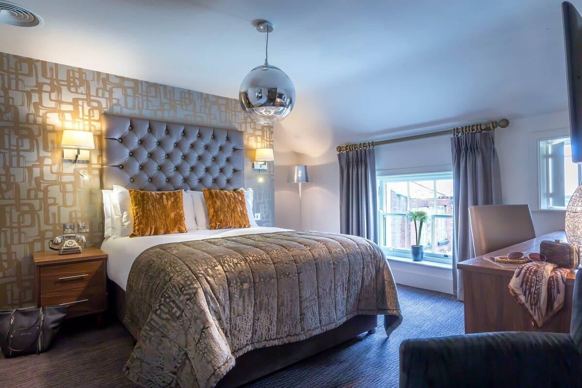 Habitación de hotel con cabecera capitonada, una cama grande con ropa de cama estampada, una lámpara de noche, un teléfono antiguo, un escritorio con lámpara y vistas a la ciudad a través de la ventana.