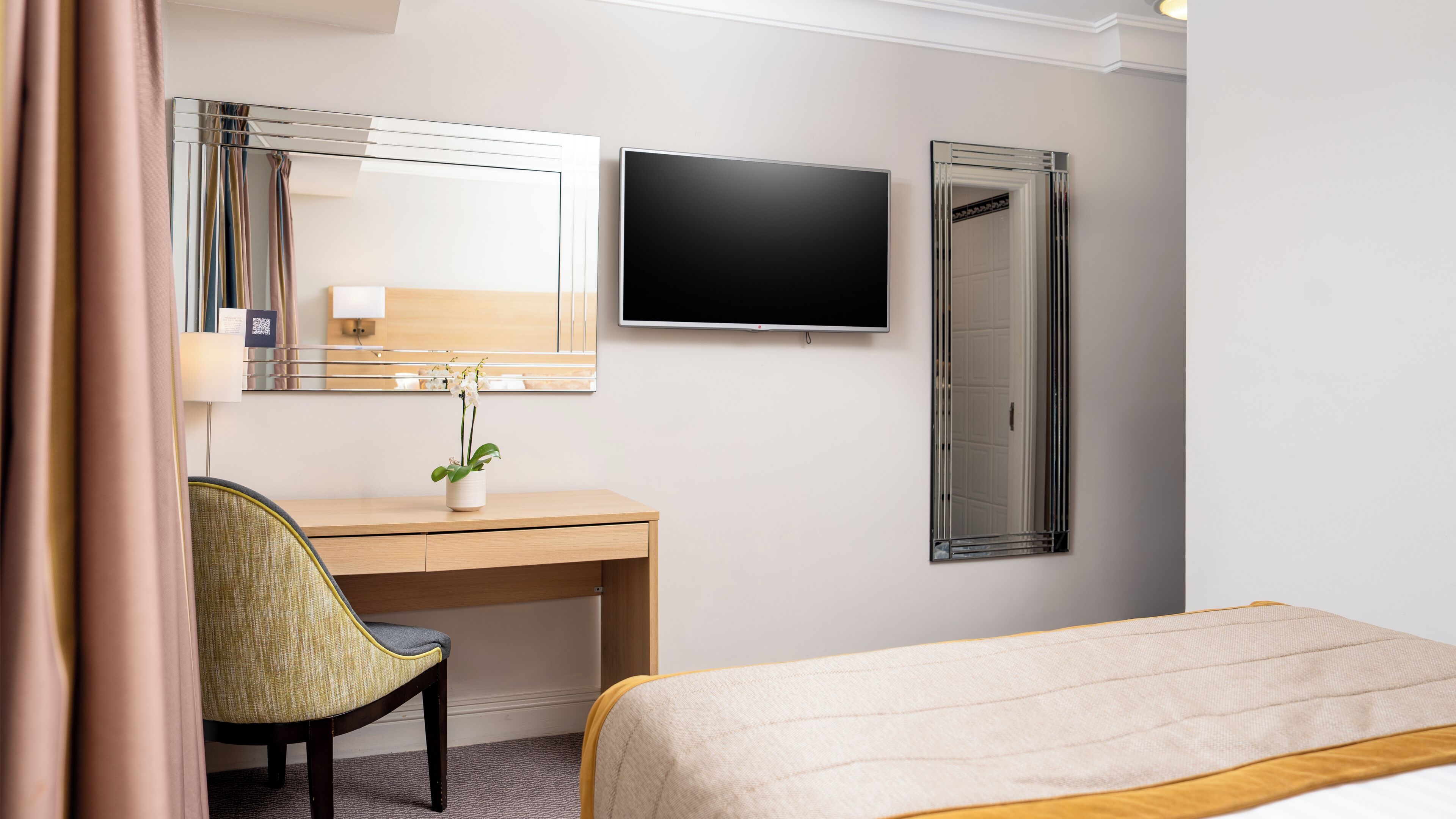 Habitación de hotel con cama, escritorio con silla, televisor de pantalla plana, espejo y planta en maceta.