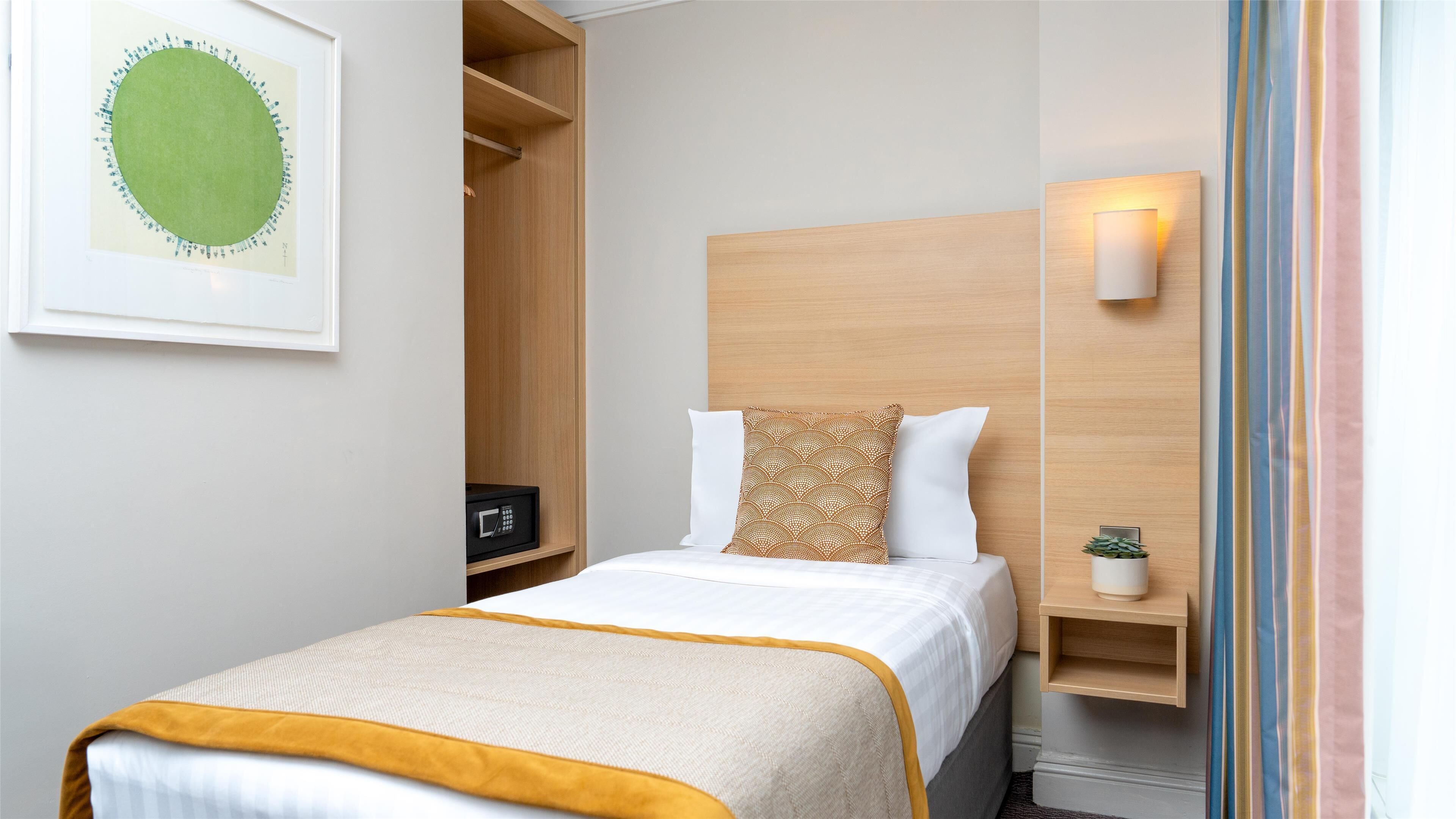 Ein ordentlich eingerichtetes Hotelzimmer mit Bett, Nachttisch, Wandlampe, gerahmtem Kunstwerk und Einbauschrank.