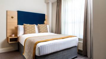 A neatly made bed with a blue headboard, two decorative pillows, a bedside table with a lamp, and a window with curtains.