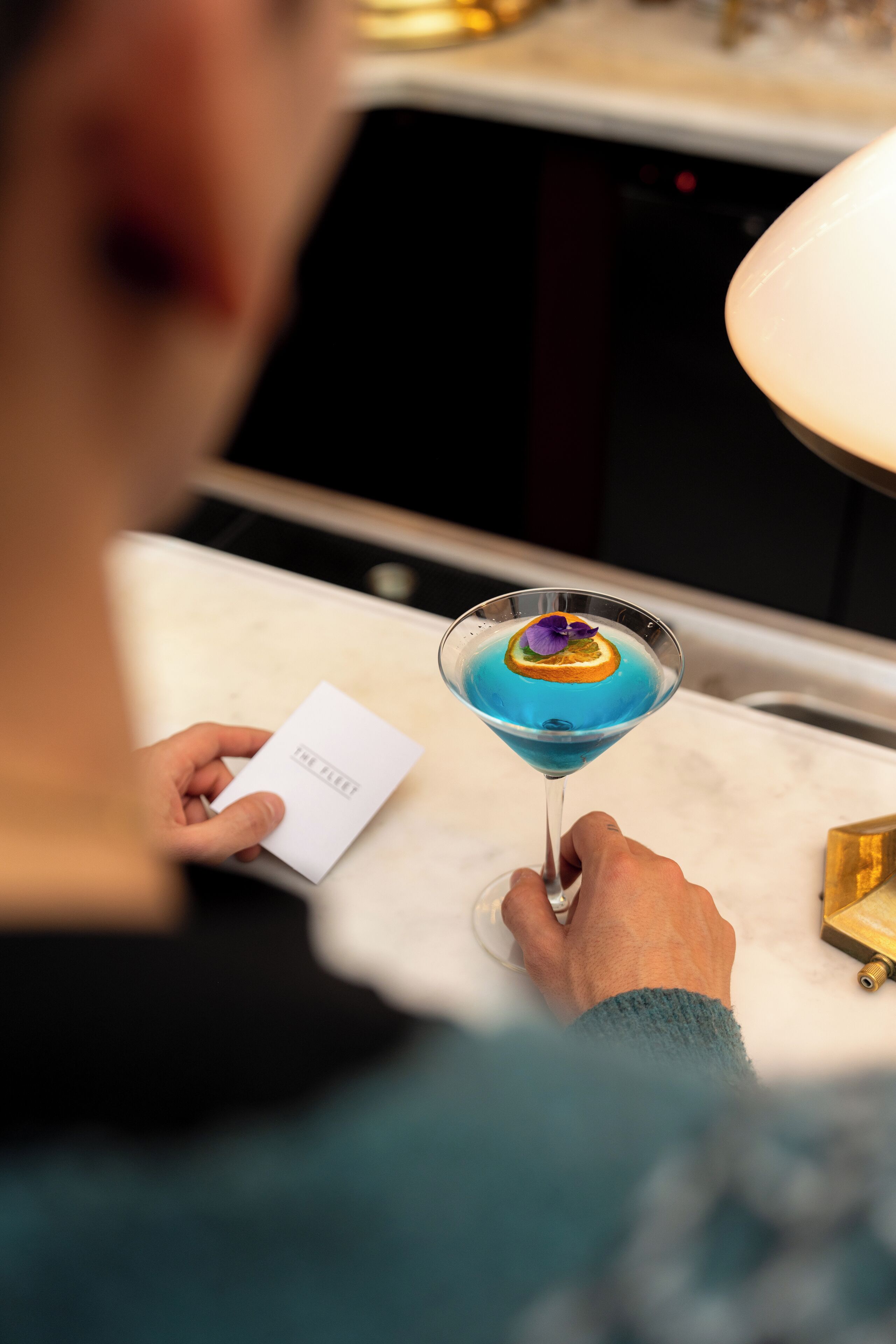 Eine Person hält ein Cocktailglas mit blauer Flüssigkeit und einer Dekoration, sitzt an einem Tisch mit einer weißen Servette und einer Speisekarte.