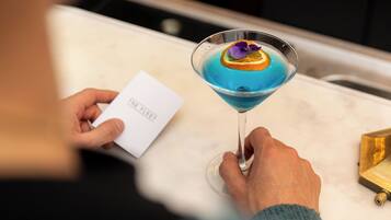 Eine Person hält ein Cocktailglas mit blauer Flüssigkeit und einer Dekoration, sitzt an einem Tisch mit einer weißen Servette und einer Speisekarte.