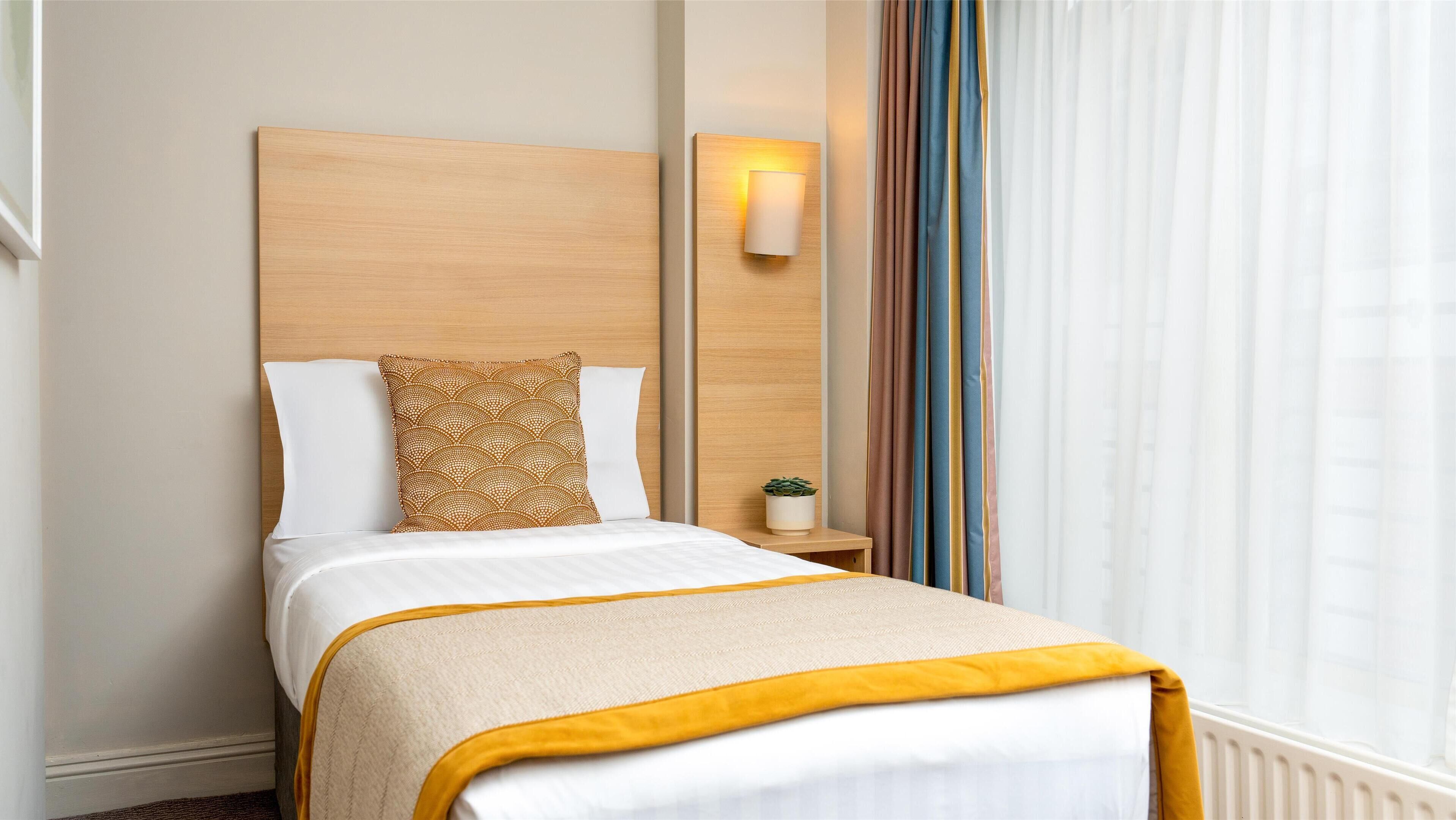 Cama bem arrumada com edredom branco e amarelo, travesseiro estampado e cabeceira de madeira. Apresenta uma mesa de cabeceira com uma planta pequena e uma luminária fixada na parede.