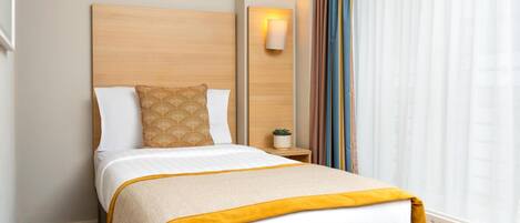Cama bem arrumada com edredom branco e amarelo, travesseiro estampado e cabeceira de madeira. Apresenta uma mesa de cabeceira com uma planta pequena e uma luminária fixada na parede.