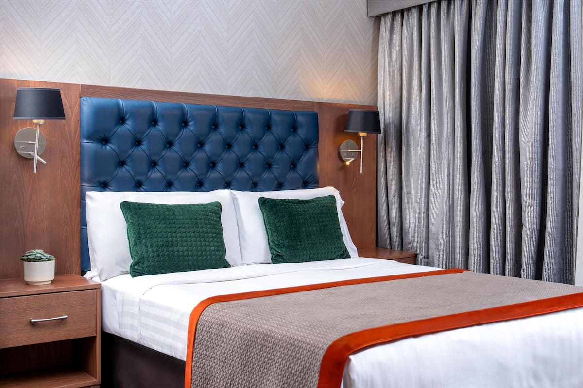 Habitación de hotel con cabecera capitonada, dos almohadas verdes, una colcha blanca, un plaid gris y naranja, y una mesita de noche con lámpara y planta en maceta.