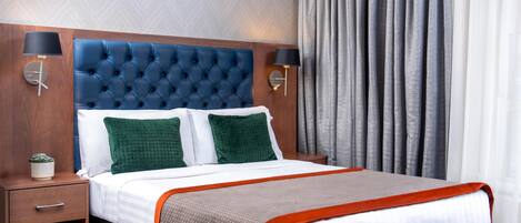 Camera d'albergo con testiera trapuntata, due cuscini verdi, copripiumino bianco, coperta grigio-arancione e comodino con lampada e pianta in vaso.