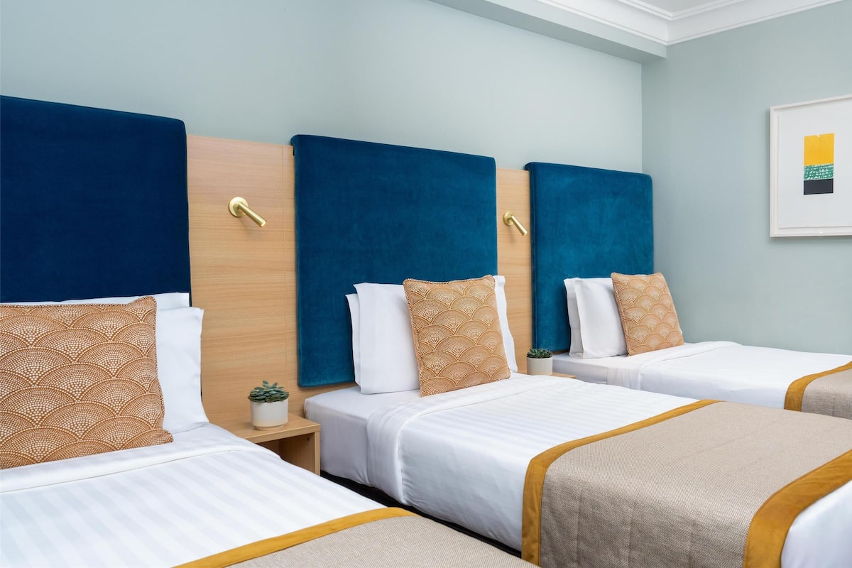 Una habitación de hotel con tres camas individuales, cada una con cabecera, mesitas de noche y almohadas decorativas.