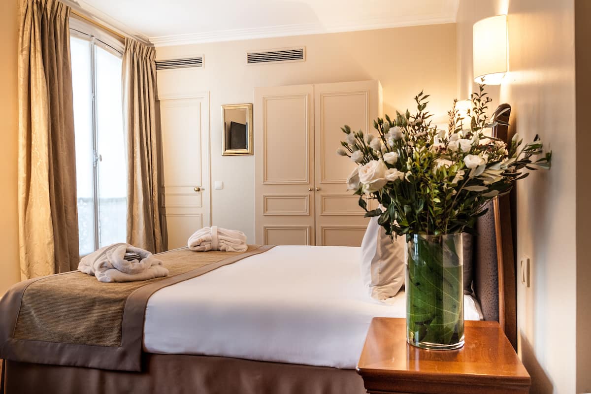 Une chambre d’hôtel avec un lit bien fait, un vase de fleurs sur une table de chevet et une fenêtre avec des rideaux.