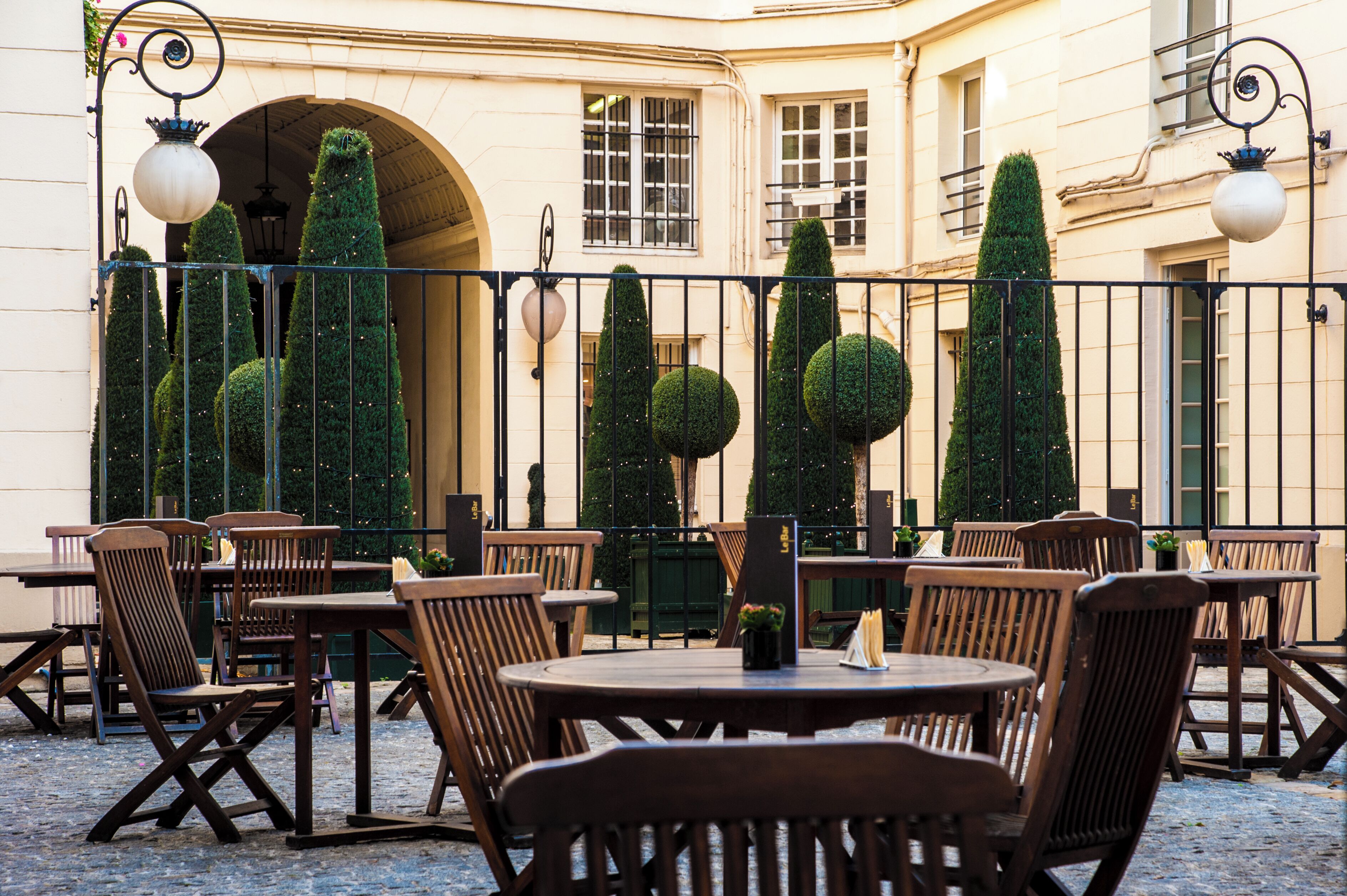 An outdoor cafe with round tables and wooden chairs, surrounded by a low fence and tall, neatly trimmed trees.