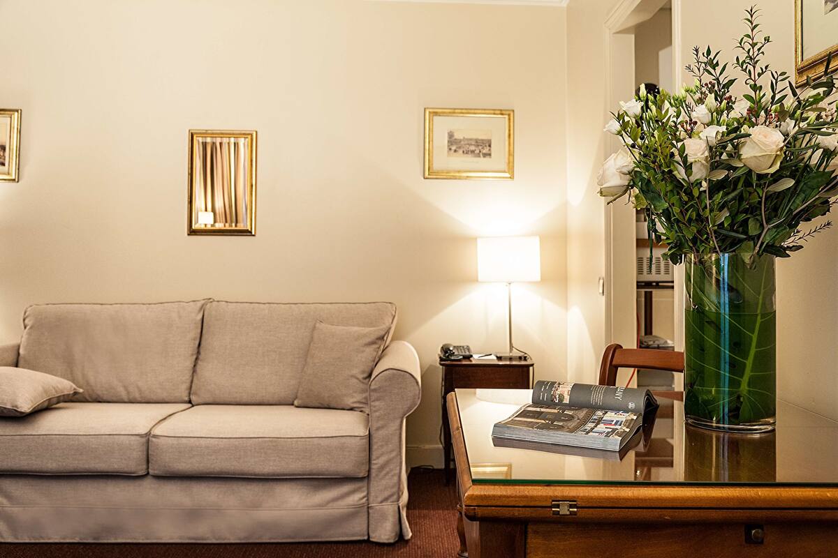 Un salon avec un canapé beige, une table basse en verre, une lampe et un vase rempli de fleurs.