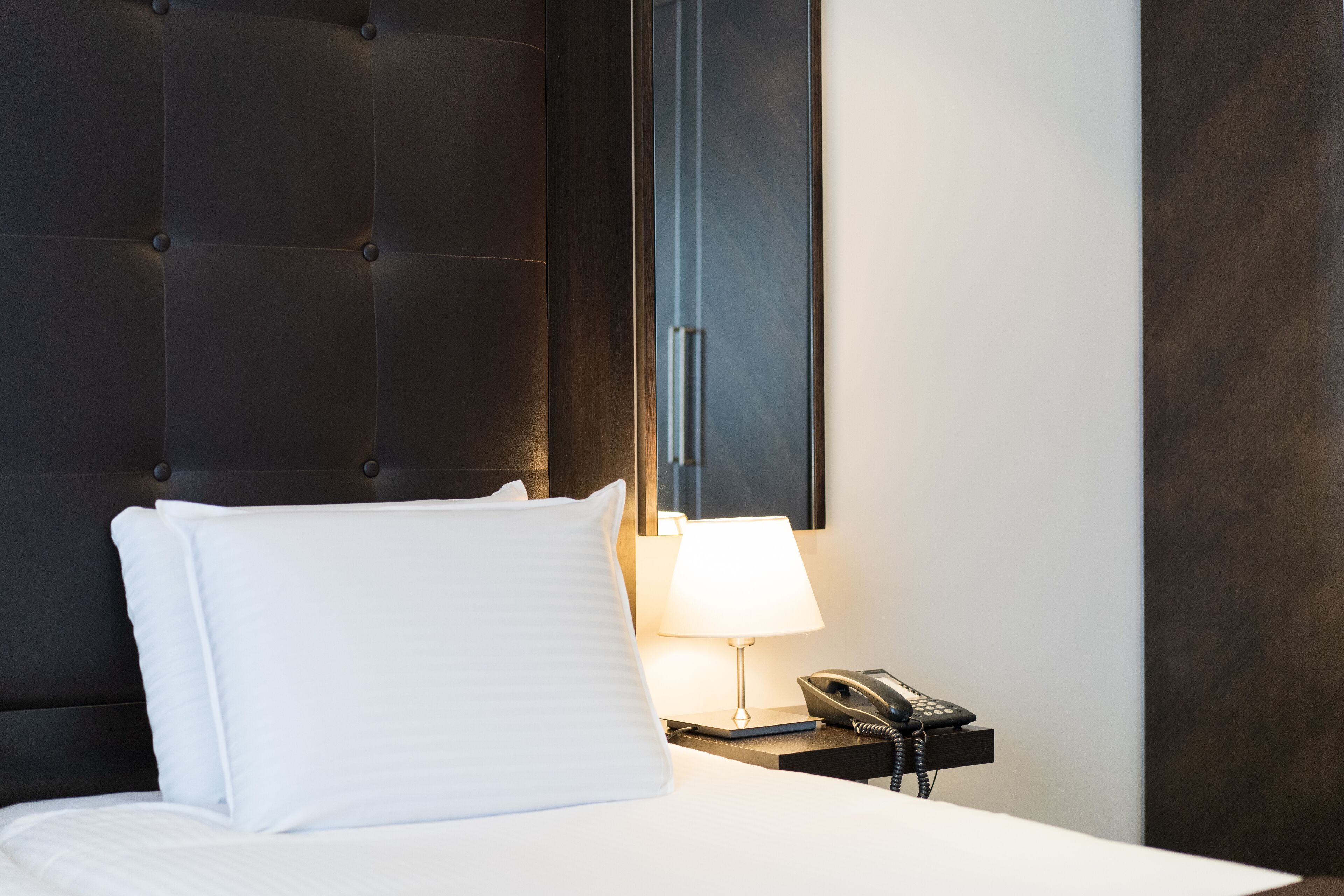 A hotel room with a tufted headboard, a white pillow, a bedside lamp, and a telephone on a nightstand.