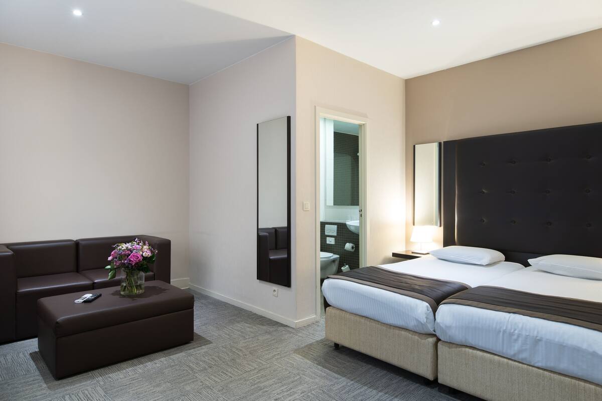 Una habitación de hotel con sofá, una mesita con un jarrón de flores, una cama con dos almohadas y un espejo grande.