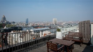 A rooftop terrace with a wooden table and chairs, a potted plant, and a view of a cityscape including a distinctive tower. - Hotel Chambord (Brussels)