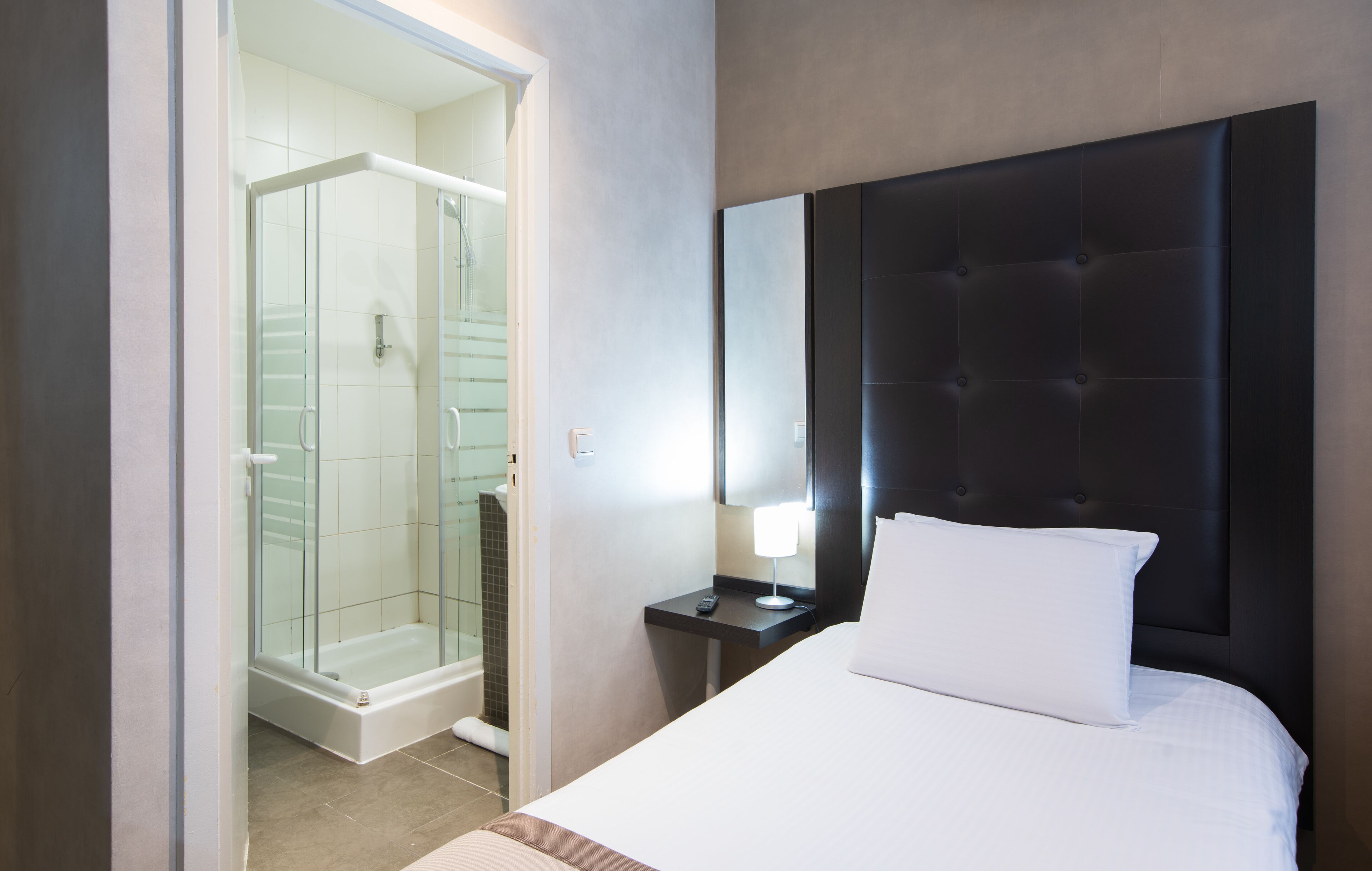 Quarto de hotel com cama, mesa de cabeceira, luminária, cabeceira e chuveiro em cabine de vidro.