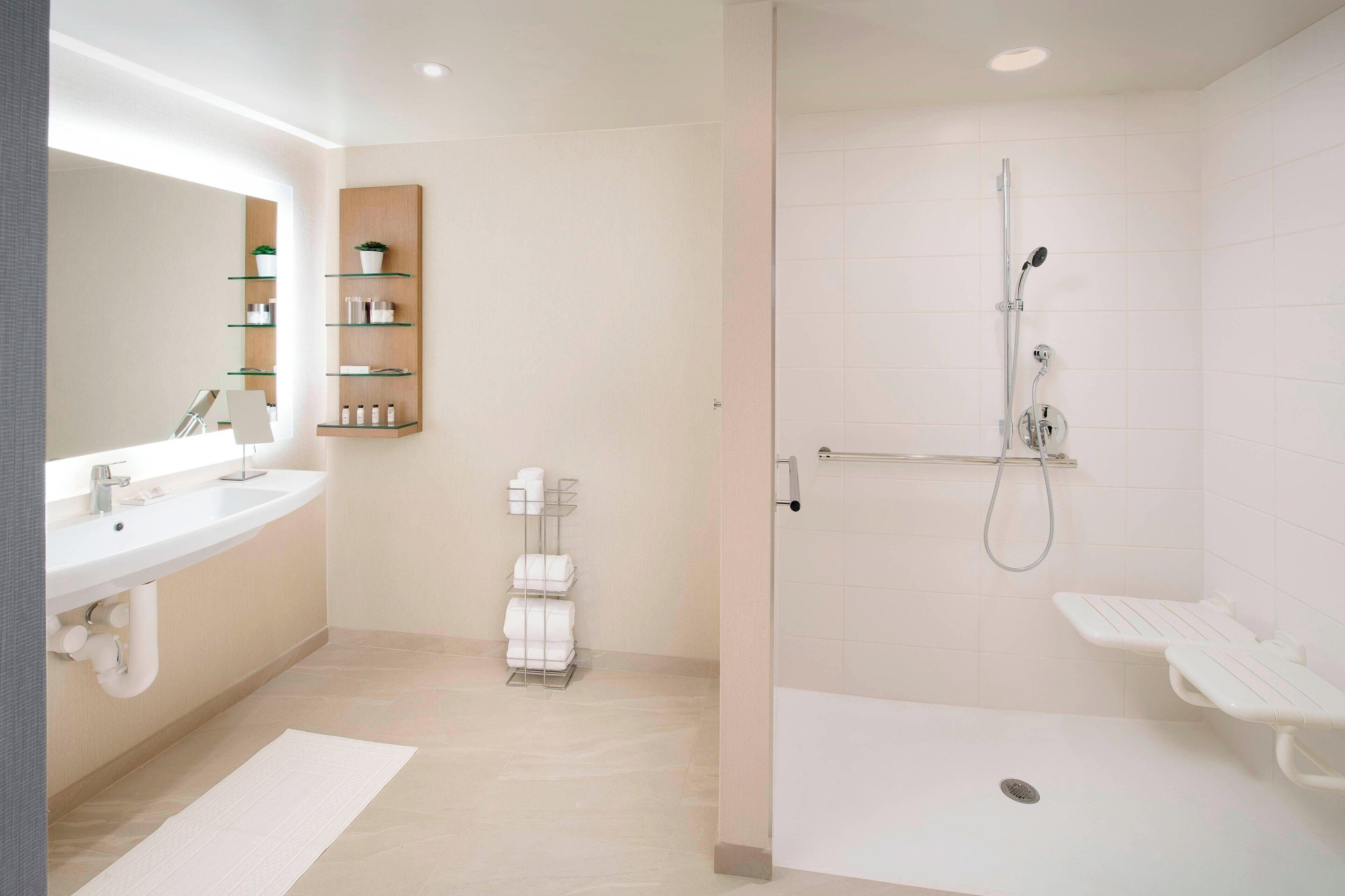 A modern bathroom with a double sink, a glass shelf unit, a shower area with a handheld showerhead, and a white bath mat.