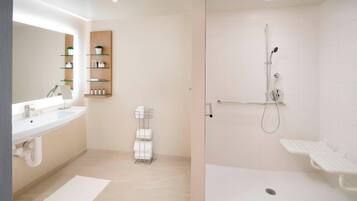 A modern bathroom with a double sink, a glass shelf unit, a shower area with a handheld showerhead, and a white bath mat.