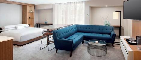 Habitación de hotel con cama, sofá azul tipo seccional, una pequeña mesa redonda, escritorio y televisión.