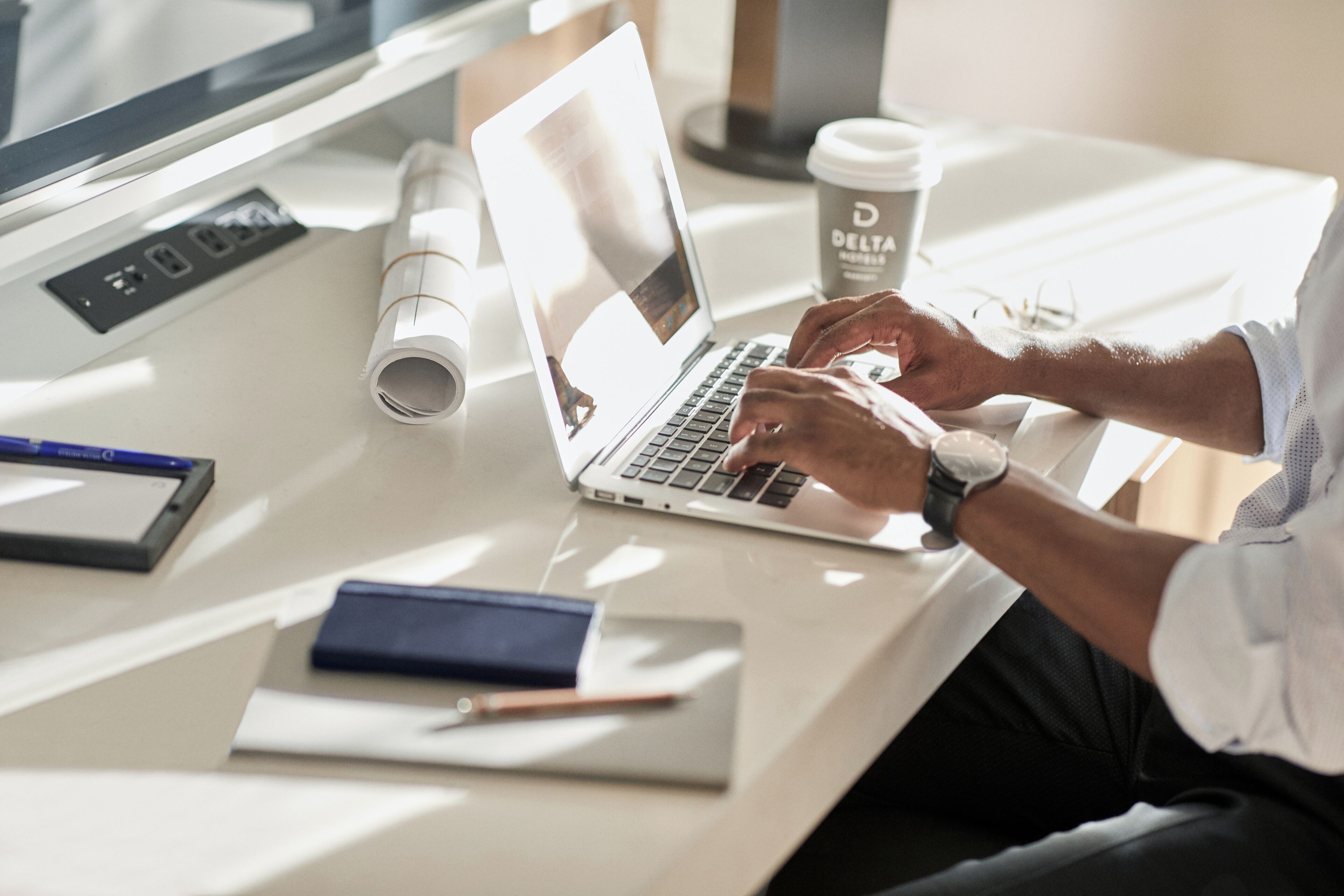 A person using a laptop on a desk with a coffee cup, rolled-up papers, and a notebook.