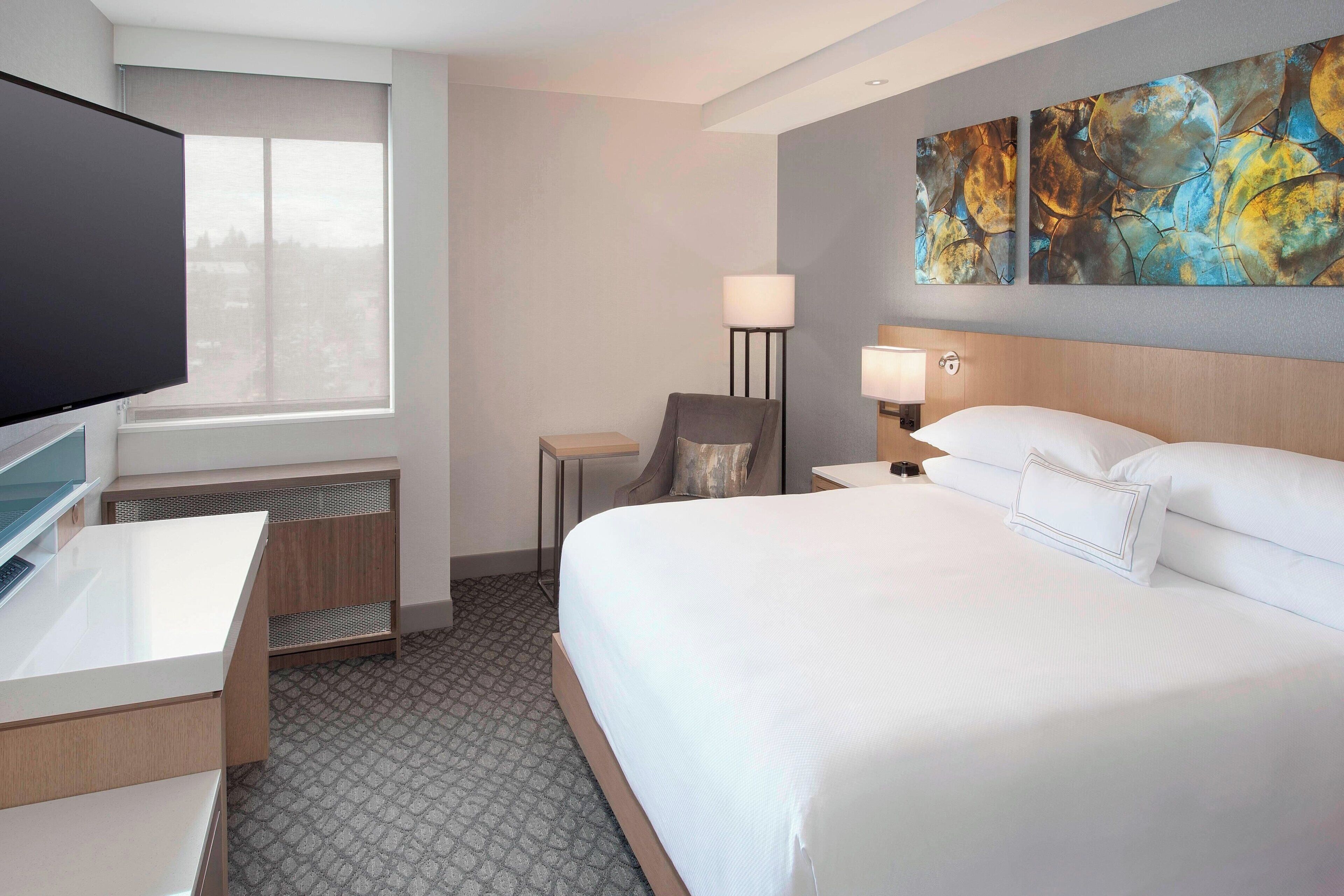 A hotel room with a bed, a desk, a chair, a TV, and artwork on the wall.