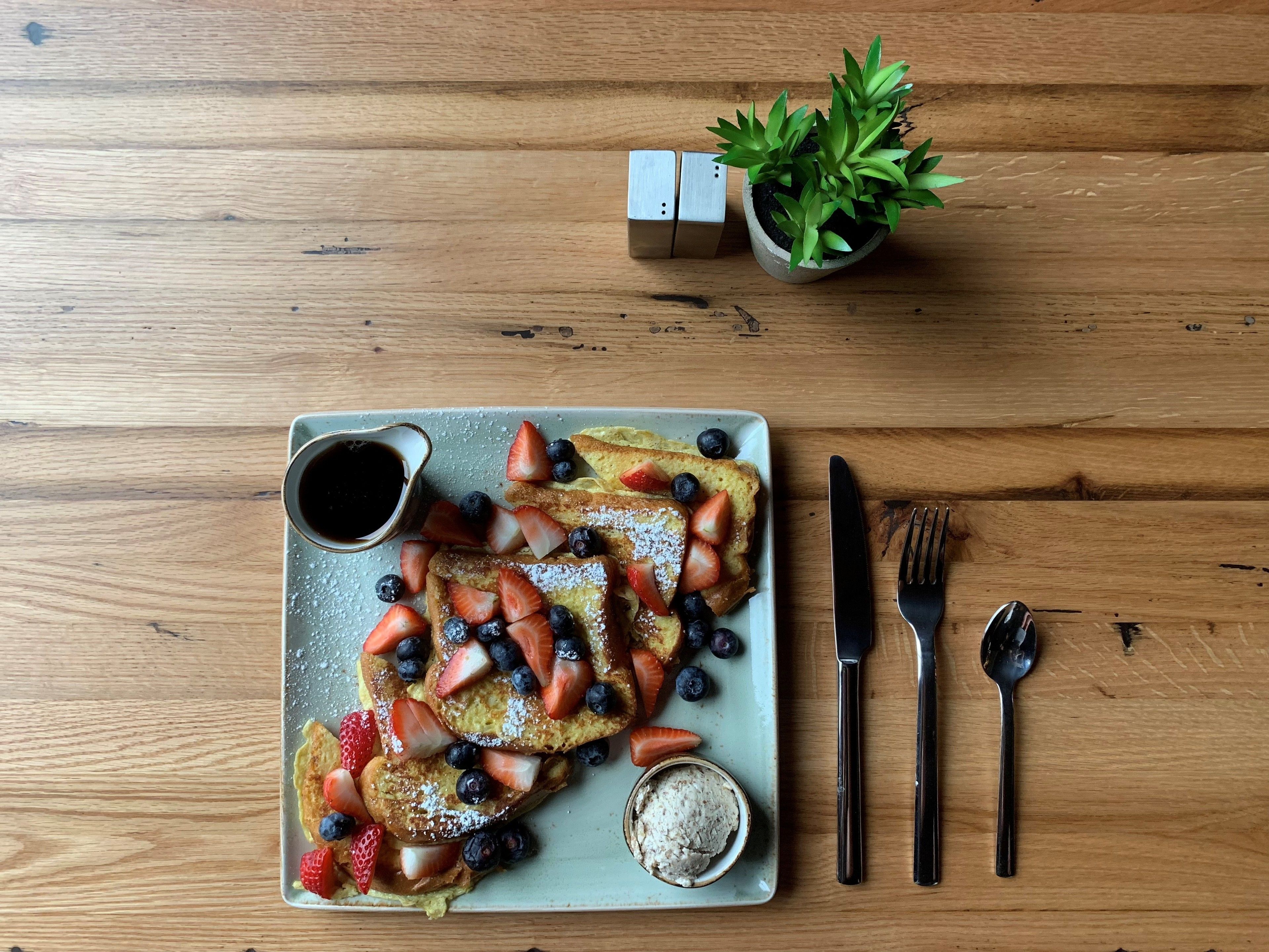 A breakfast plate with French toast, strawberries, blueberries, and powdered sugar, accompanied by a small bowl of ice cream and a cup of syrup, on a wooden table.