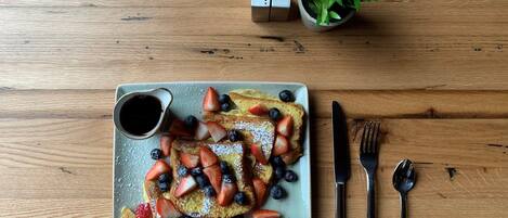 Un plato de desayuno con tostadas francesas, fresas, arándanos y azúcar glas, acompañado de un pequeño recipiente de helado y una taza de jarabe, sobre una mesa de madera.