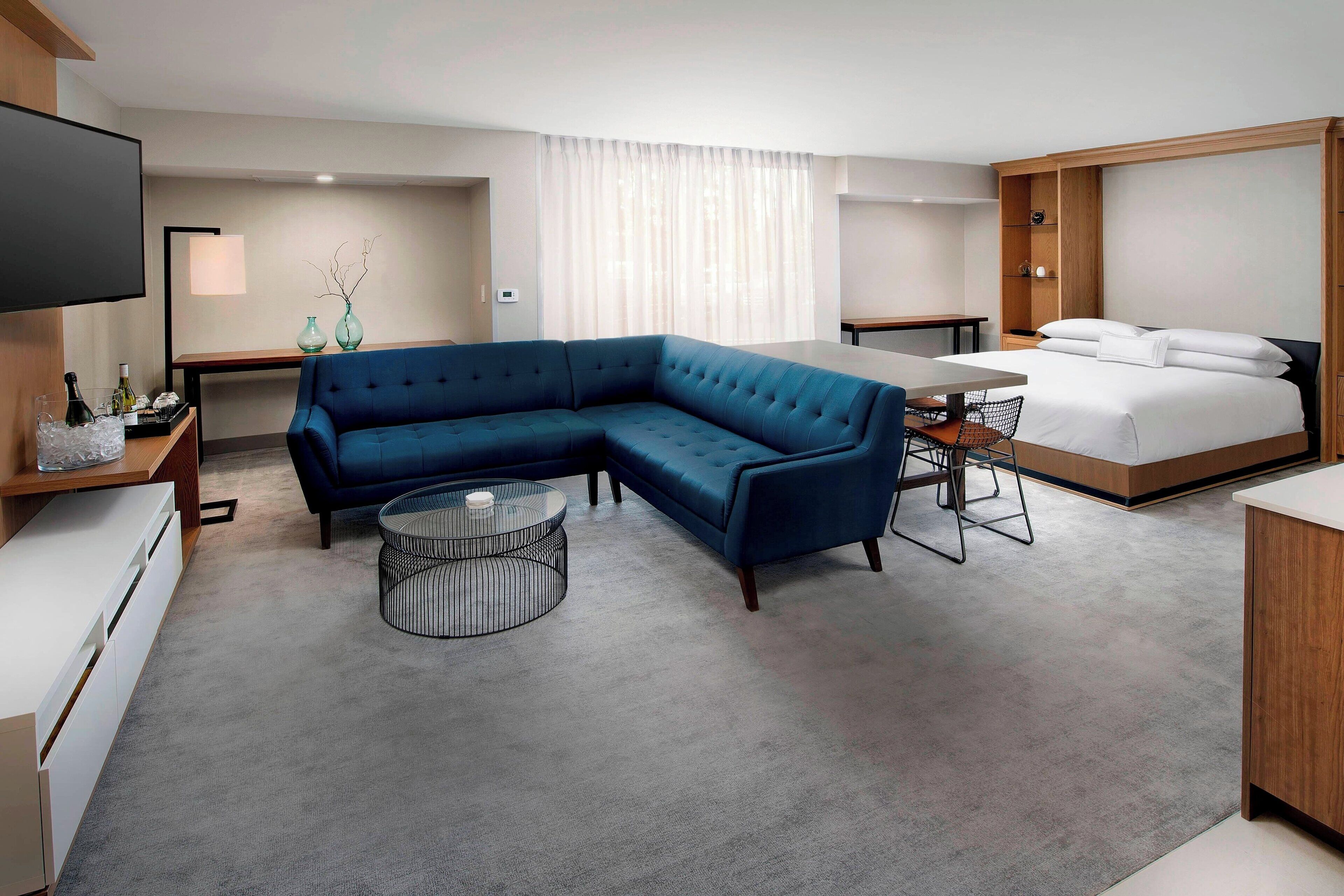 A modern hotel room with a sectional sofa, a round coffee table, a bed with white linens, a desk with a computer, and a flat-screen TV mounted on the wall.