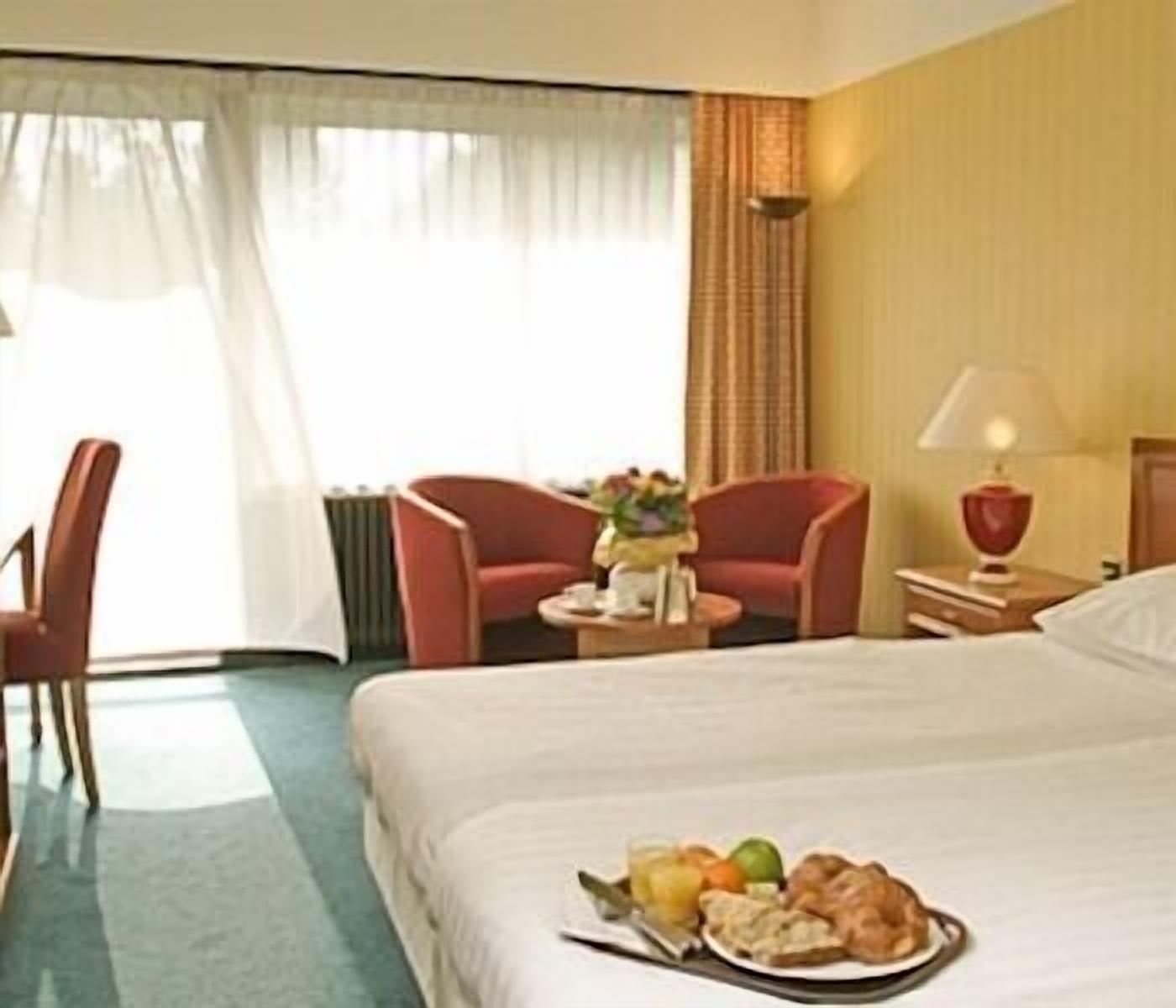 Habitación de hotel con una cama, un escritorio con lámpara, una silla y una pequeña mesa con un plato de comida.