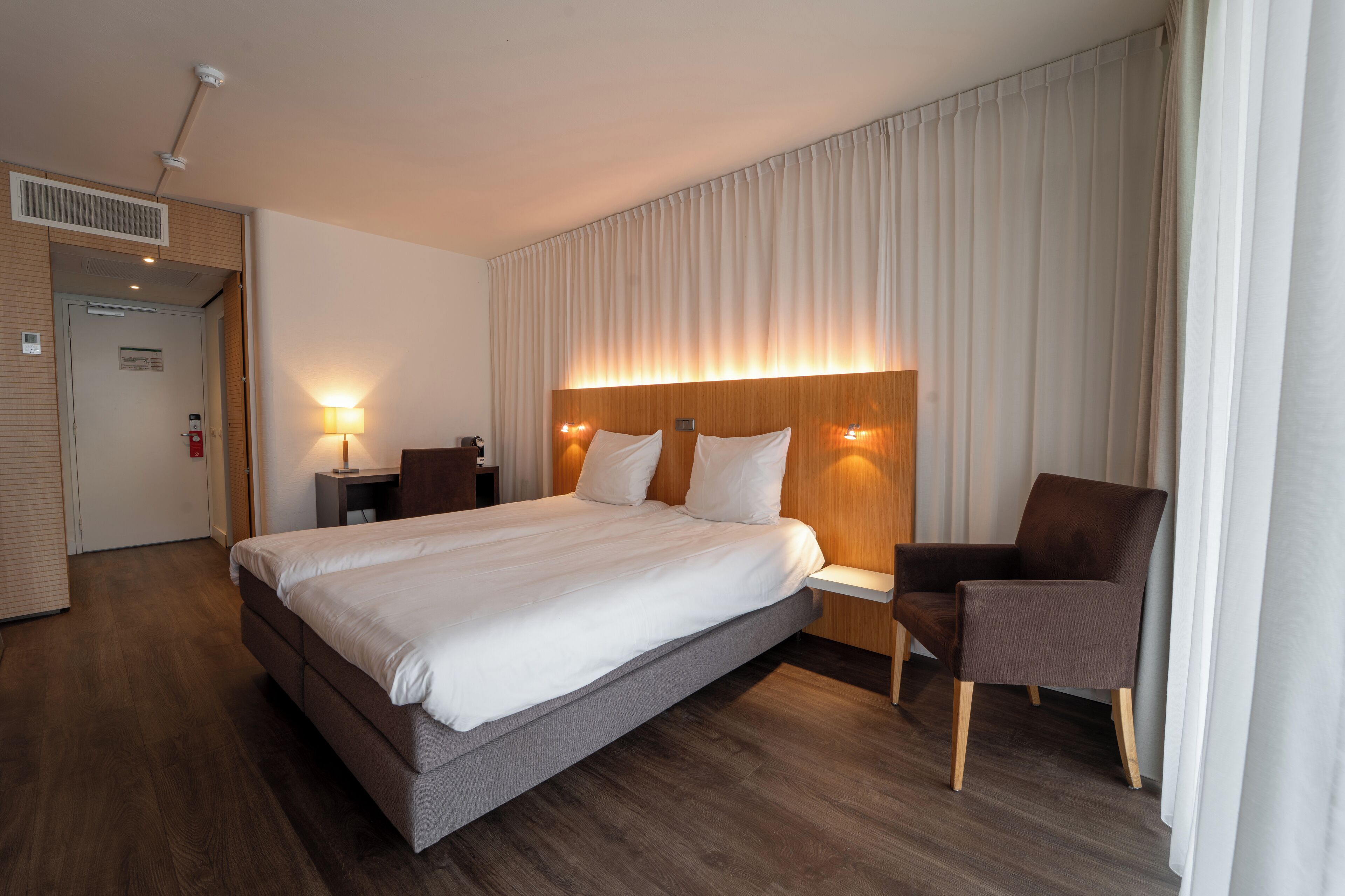 Una habitació d'hotel moderna amb un llit gran, un panell de capçalera de fusta, una petita taula de nit, una cadira i una finestra amb cortines.
