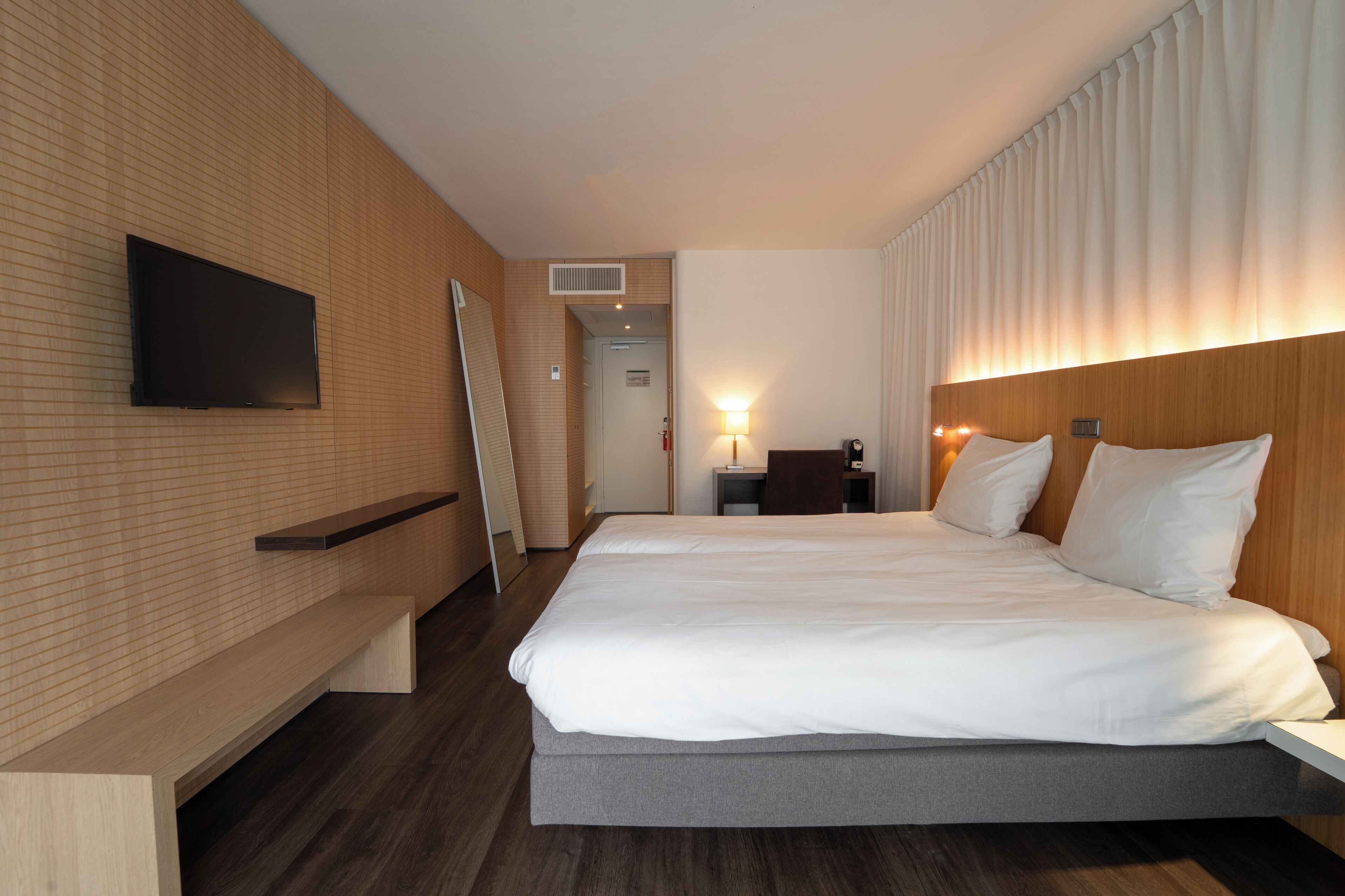 Una habitación de hotel moderna con una cama grande, un televisor de pantalla plana montado en la pared, un escritorio con una silla y un banco a lo largo de la pared.