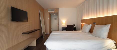 Una habitación de hotel moderna con una cama grande, un televisor de pantalla plana montado en la pared, un escritorio con una silla y un banco a lo largo de la pared.