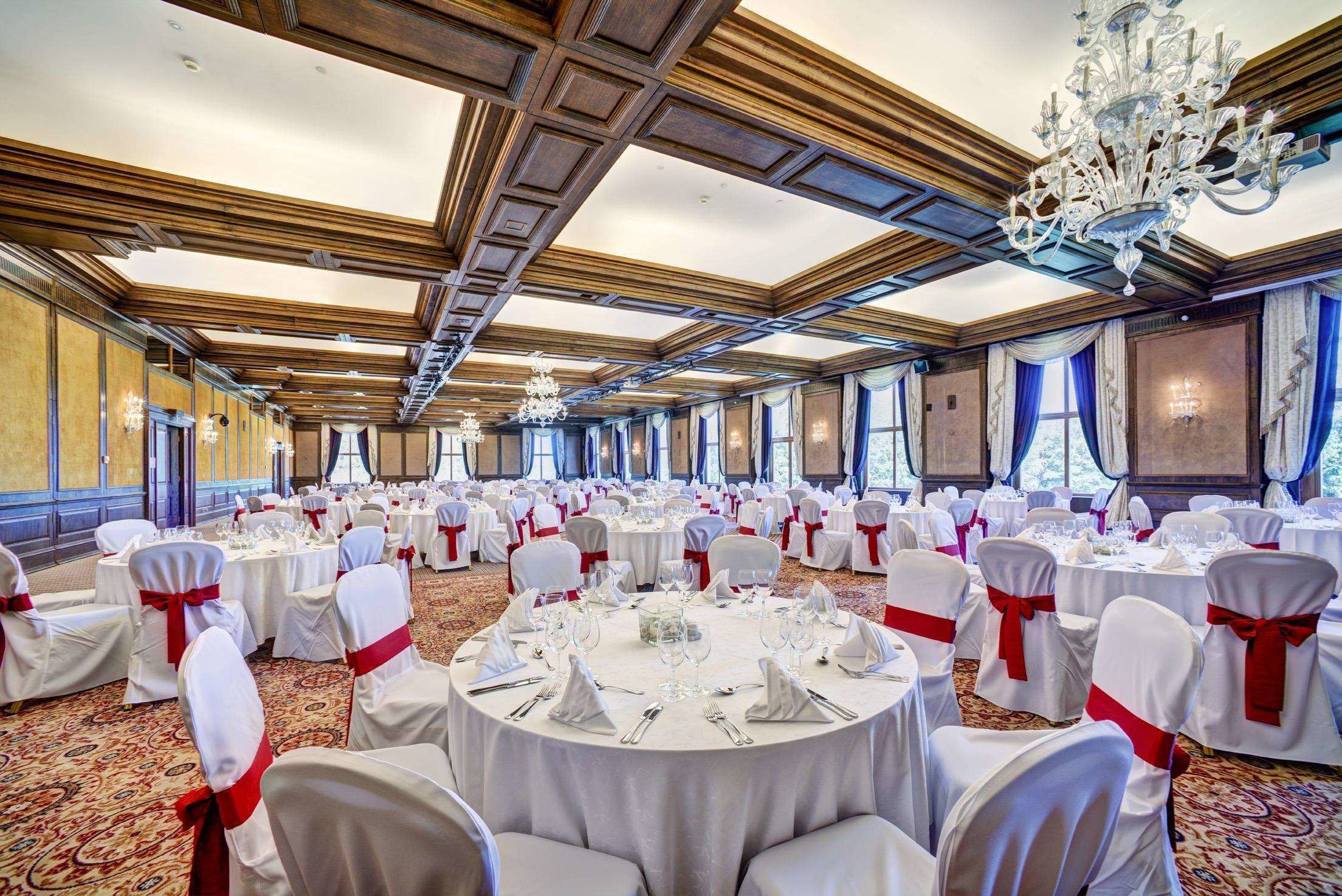 Een balzaal met ronde tafels gedekt voor een formeel evenement, met kroonluchters en een hoog plafond met houten lambrisering.