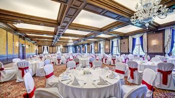 Een balzaal met ronde tafels gedekt voor een formeel evenement, met kroonluchters en een hoog plafond met houten lambrisering.