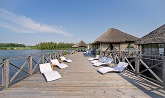 Un muelle de madera con sillones de descanso y construcciones de techo de paja que da a un lago.