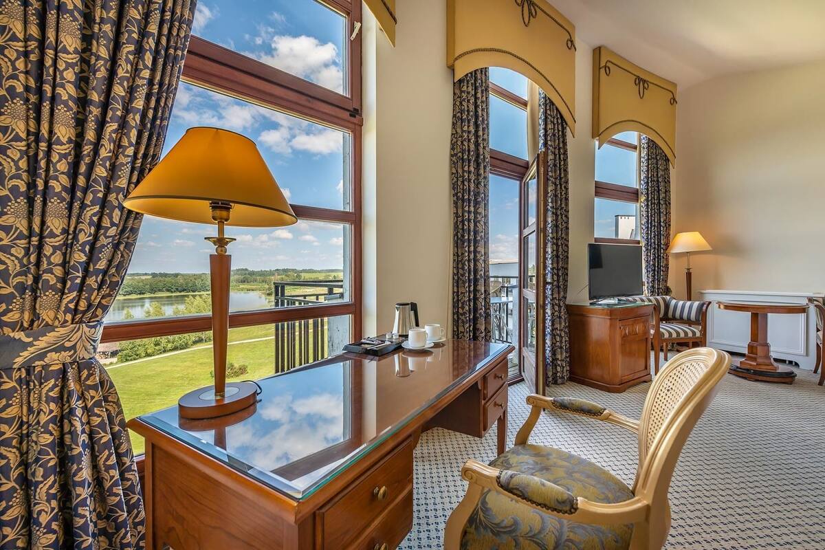 A hotel room with a large window offering a view of a lake and greenery, a desk with a lamp, a television, and a seating area with a chair and a small table.