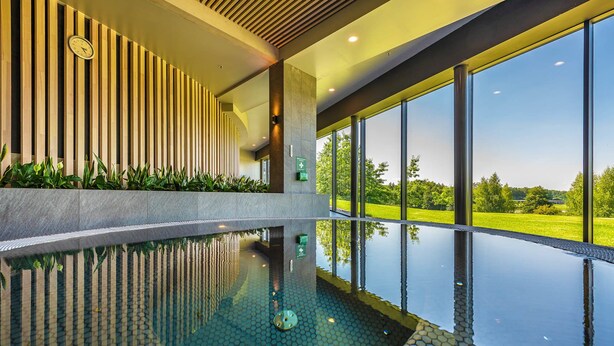 A modern indoor swimming pool with a hexagonal tile pattern, large windows offering a view of a green landscape, and a wooden slatted wall design. - Vilnius Grand Resort (Kalikstiškes)