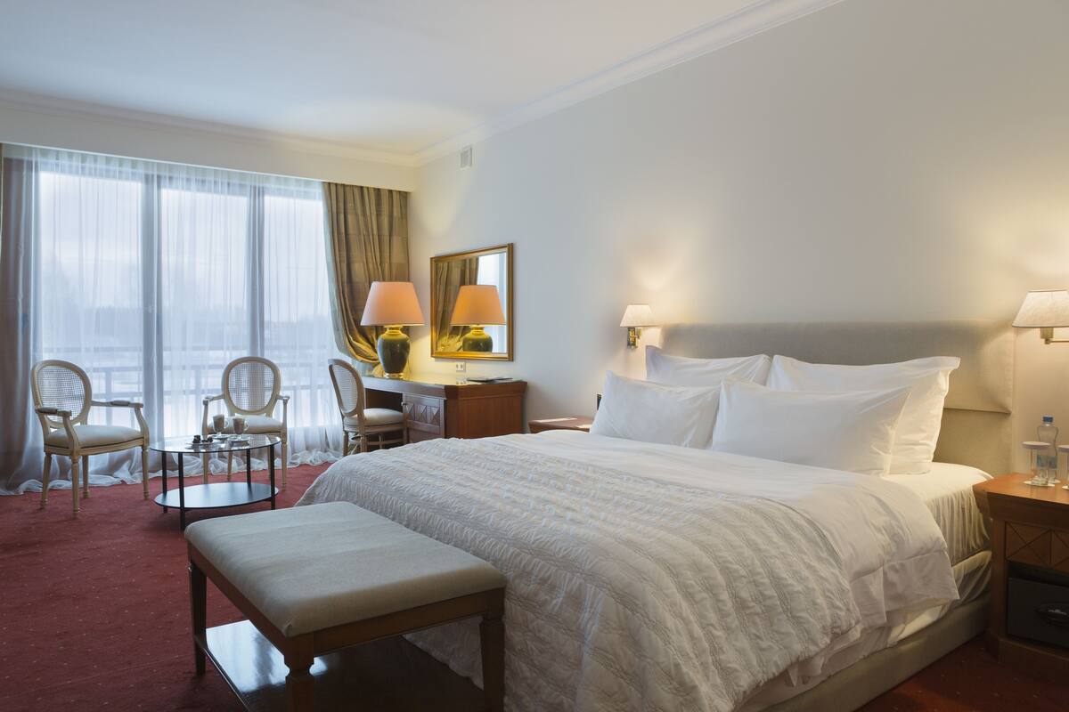 A hotel room with a large bed, two bedside tables, a small round table, two chairs, a bench, and a large window with curtains.
