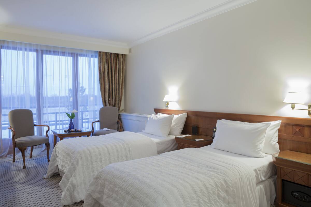 A hotel room with two beds, a chair, a small table with a vase, and a nightstand with a lamp.