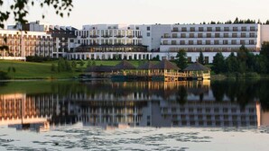 A modern hotel with multiple floors, a reflection in a calm lake, and a grassy area with a gazebo. - Vilnius Grand Resort (Kalikstiškes)