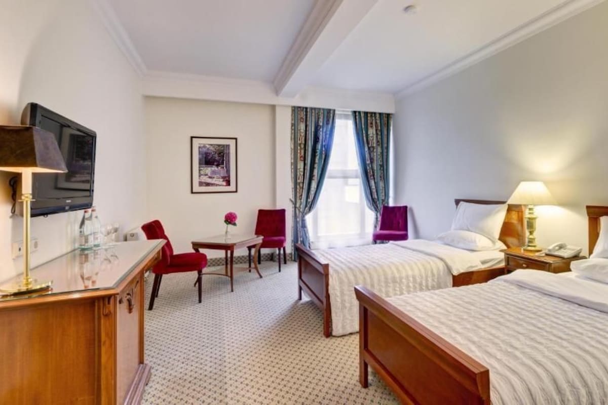 A hotel room with two beds, a desk, a chair, a TV, and a window with curtains.