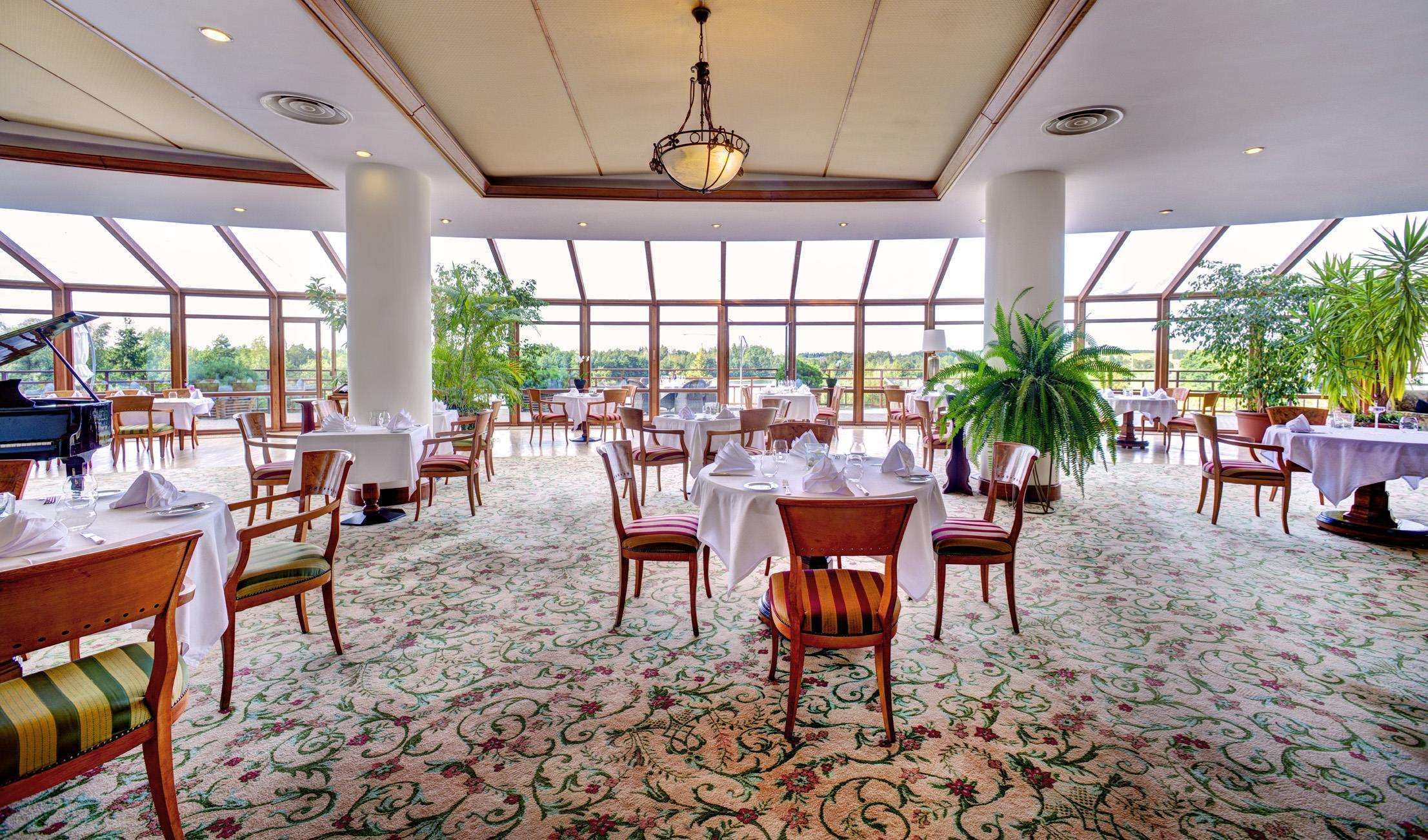 A spacious dining area with tables set for guests, large windows offering a view of the outdoors, and a grand piano in the corner.