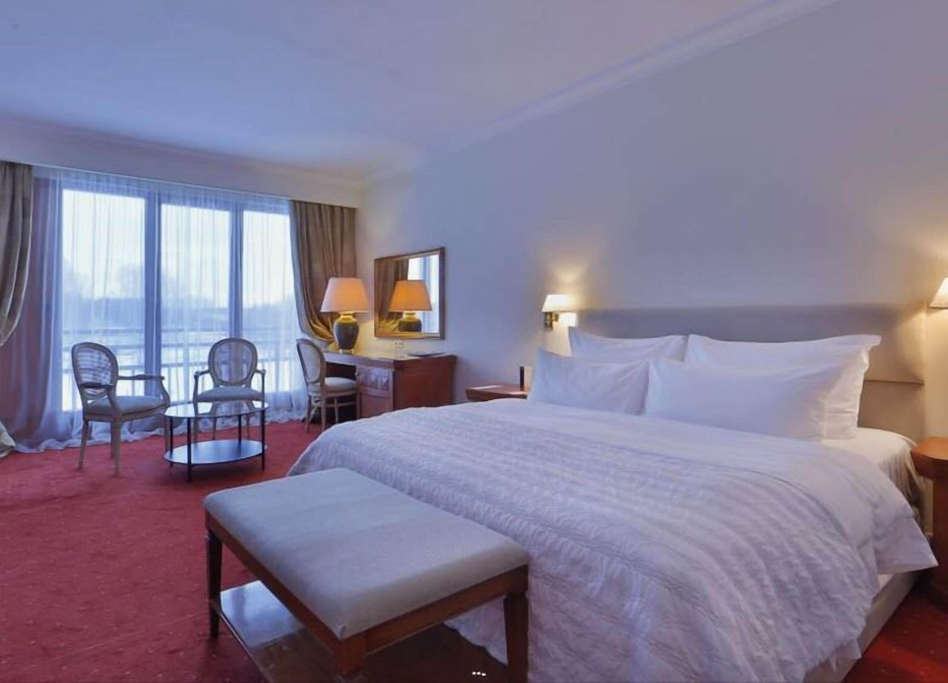 Habitación de hotel con una cama grande, un escritorio con dos sillas, una ventana con cortinas y vistas a la ciudad.
